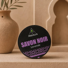 Amalia Savon Noir Lavande soap bar on a beige surface with a decorative vase and plant in the background.